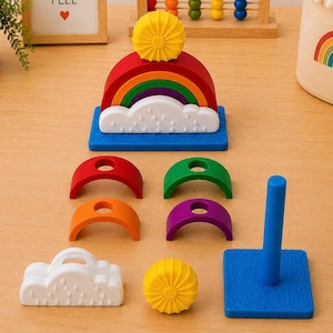 May include: A rainbow stacking toy featuring a blue base with a central post, a white cloud piece with embossed raindrops, and colorful arched segments in red, orange, green, and purple. A yellow sun-shaped piece sits atop the stack.