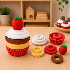 May include: A stackable cupcake toy composed of modular pieces including a red strawberry topper, white frosting layer, brown chocolate ring, yellow middle section, and a red fluted base. Additional disassembled pieces are arranged nearby.