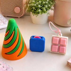 以下が含まれることがあります： カラフルなフィジェットおもちゃのコレクション。緑とオレンジ色の円錐形のおもちゃ、ピンクのローラーが付いた青い立方体、4つのピンクのボタンが付いた白とピンクの正方形が表示されています。