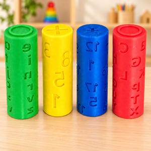 May include: Four cylindrical rollers in green, yellow, blue, and red, each featuring embossed letters, numbers, or mathematical symbols on their surfaces. These plastic tools are designed for imprinting patterns into soft materials like clay or dough.