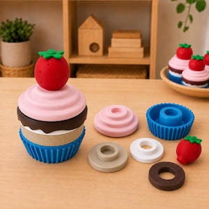 May include: A stackable cupcake toy composed of modular pieces including a blue fluted base, a tan ring, a brown ring, a pink top, and a red strawberry topper. Additional loose pieces include a pink disc, a blue base, a white ring, and a brown ring.