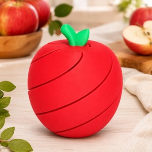 May include: A red apple-shaped kitchen gadget with a green stem and leaf design. The apple has a segmented design, and is sitting on a light-coloured surface. Apples and a wooden bowl are in the background.