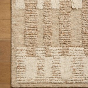 Puede incluir: Una alfombra tejida con un patrón geométrico de secciones alternas de lana copetuda de pelo largo color crema y fibras de yute beige tejidas planas. El diseño consiste en bloques rectangulares verticales y horizontales de textura.