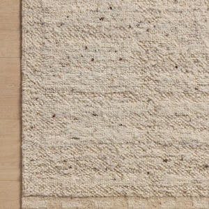 May include: A rectangular area rug featuring a thick, cream-colored textured pile with scattered brown and grey flecks throughout. The bottom edge displays a distinct horizontal border with a repeating light tan geometric pattern.