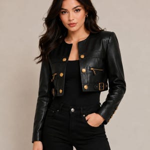 May include: A black cropped jacket featuring a round neckline, long sleeves, and a front button closure. The jacket is adorned with gold-toned buttons, two horizontal zippered pockets on the chest, and adjustable side buckles at the waistline.