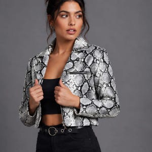 Può includere: Una giacca corta con motivo a pelle di serpente bianco e nero, colletto a punta, maniche lunghe e una tasca orizzontale con cerniera sul petto sinistro. La giacca è indossata sopra un top nero semplice e pantaloni neri con cintura argentata.