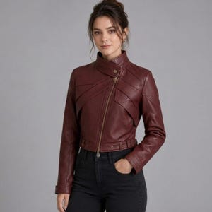 Peut inclure: Veste en cuir bordeaux avec fermeture éclair asymétrique et taille ceinturée. La veste présente un col montant et des détails matelassés sur les épaules. Le modèle porte un jean noir.