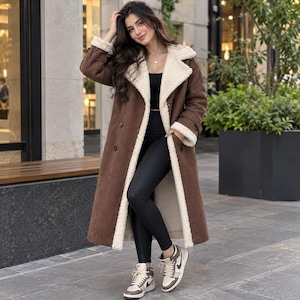 May include: A long brown suede coat with a cream-colored faux shearling lining and collar, worn over a black top and black leggings, paired with high-top sneakers featuring brown, white, and black color blocking and white laces.