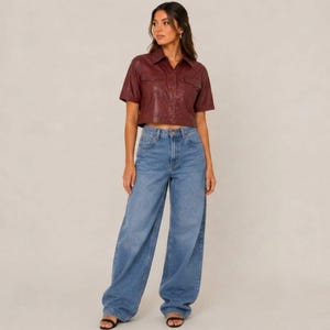 May include: A woman in a burgundy short-sleeved collared shirt and light blue wide-leg jeans. The shirt has two front pockets. The jeans are high-waisted and have a relaxed fit. The woman is wearing black sandals.