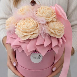 Op de afbeelding: Een ronde roze geschenkdoos met crèmekleurige bloemvormige items, geaccentueerd met roze tule en een roze satijnen lint. Een rond wit label op de voorkant van de doos bevat de tekst Touch of Scent met een kleine krulafbeelding.