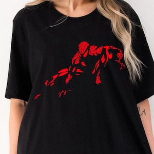 May include: A black short-sleeved t-shirt featuring a bold, red graphic print of a superhero character in a crouching, web-slinging pose. The design is centered on the chest, showcasing a detailed web pattern across the character's suit.