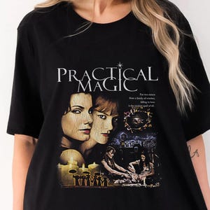 Puede incluir: Una camiseta negra de manga corta con un estampado gráfico que incluye el texto "PRACTICAL MAGIC" y "For two sisters from a family of witches, falling in love is the trickiest spell of all." El diseño muestra retratos de dos mujeres.