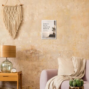 May include: A framed print with the text "FOCUS PLAN EXECUTE REPEAT" is displayed on a textured wall. The print features a minimalist design with a staircase, a small tree, and a body of water. A macrame wall hanging and a lamp are also visible.