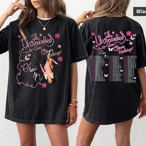 Könnte beinhalten: Ein schwarzes Kurzarm-T-Shirt mit pinkfarbenem Grafikdruck. Die Vorderseite zeigt den Text The Unraveled Tour Olivia Rodrigo mit einer Illustration einer Person auf einer Schaukel. Die Rückseite listet Tourdaten und Orte in pinkem Text.