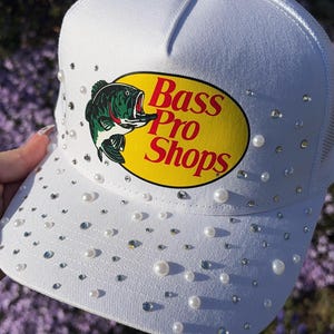 Puede incluir: Gorra de camionero blanca con el logo "Bass Pro Shops" en amarillo y rojo, con un gráfico de pez. La gorra está adornada con perlas y pedrería dispersas.