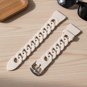 May include: Two off-white watch bands with a chain link design. One band has a silver buckle. The bands are lying on a light wood surface, with a wooden tray and sunglasses in the background.