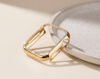 May include: A gold-colored watch case with a rectangular shape. The case is adorned with small, sparkling crystals around the edges. The watch case is designed to protect the watch face and add a touch of elegance.