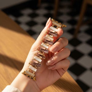 Op de afbeelding: Een goudkleurige horlogeband met rechthoekige, iriserende segmenten in wit, roze en bruin. De band wordt in een hand gehouden, waarbij het decoratieve ontwerp wordt getoond. De horlogeband heeft een decoratieve sluiting met kleine, heldere stenen.