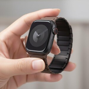 May include: A black smartwatch with a dark brown metal band is held in a hand. The watch face displays a clock design with white hands. The watch has a black case with a red button on the side.