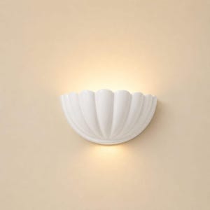 May include: A white ceramic wall light shaped like a half-shell with vertical fluted ridges. The fixture emits a warm, soft glow from its top opening, casting light upwards against the wall surface while maintaining a clean, curved profile.