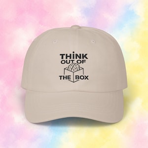 Könnte beinhalten: Eine beige Baseballkappe mit einem schwarzen Grafikdruck auf der Vorderseite. Das Design zeigt eine Strichzeichnung eines Gehirns in einer Box mit dem Text "THINK OUT OF THE BOX" in einer fetten, serifenlosen Schriftart gedruckt.