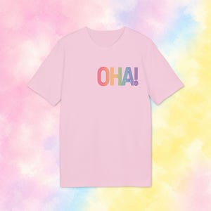 Könnte beinhalten: Ein hellrosa T-Shirt mit kurzen Ärmeln und dem Schriftzug OHA! auf der Brust. Die Buchstaben sind in einem mehrfarbigen, gestreiften Regenbogenmuster gestaltet, das aus roten, orangen, gelben, grünen, blauen und violetten Linien besteht.