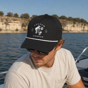 May include: Black baseball cap with white rope detail and a white graphic that reads "POSSUM KINGDOM" with a fish illustration. The hat is worn by a person on a boat, with a lake and cliffs in the background.
