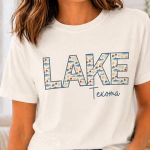 May include: Cream-colored t-shirt with the word "LAKE" in large, blue outlined letters filled with a pattern of lake-themed illustrations. Below, the word "Texoma" is written in a script font.
