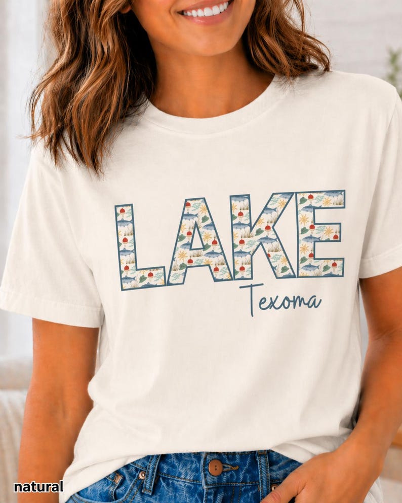 May include: Cream-colored t-shirt with the word "LAKE" in large, blue outlined letters filled with a pattern of lake-themed illustrations. Below, the word "Texoma" is written in a script font.