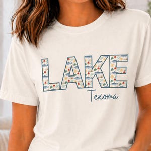 May include: Vintage white t-shirt with the word "LAKE" in large, patterned letters, and "Texoma" in a smaller, cursive font. The "LAKE" letters are filled with a design of boats, suns, and trees. The shirt is a casual, short-sleeved style.