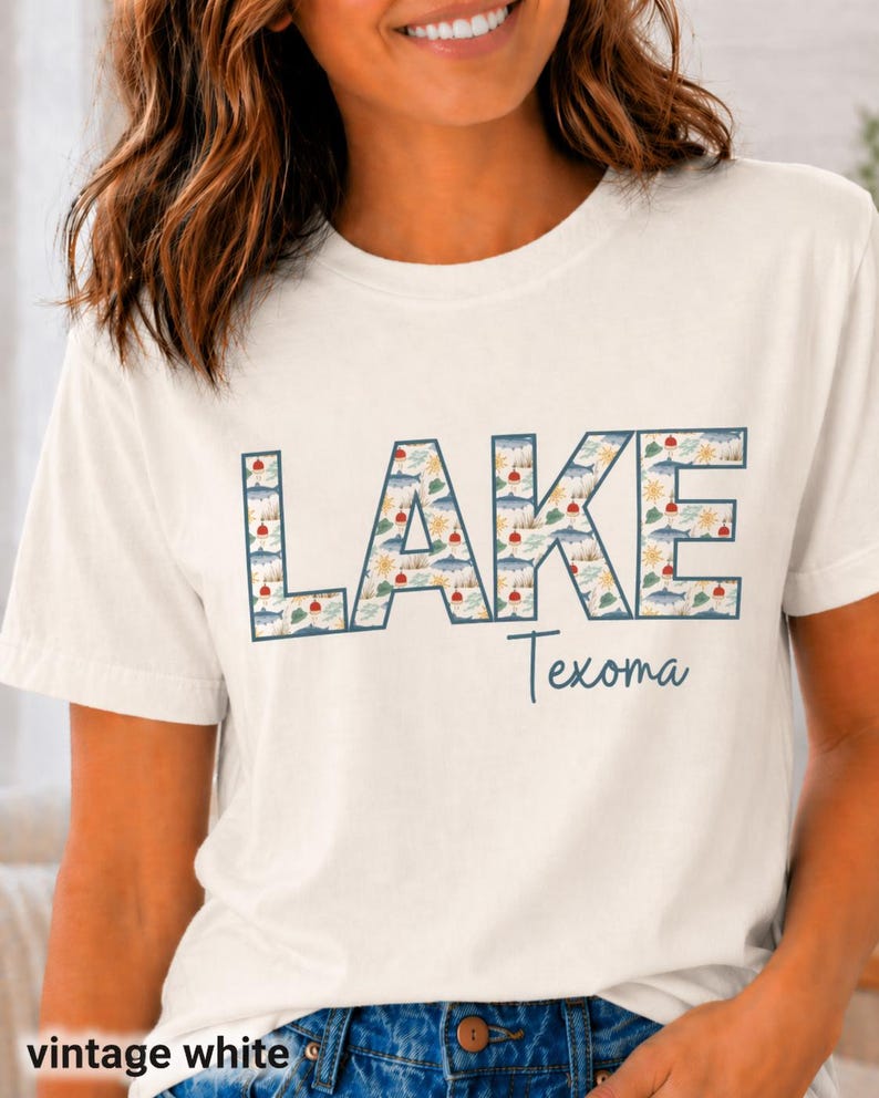 May include: Vintage white t-shirt with the word "LAKE" in large, patterned letters, and "Texoma" in a smaller, cursive font. The "LAKE" letters are filled with a design of boats, suns, and trees. The shirt is a casual, short-sleeved style.