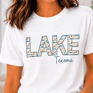 May include: White t-shirt with the word "LAKE" in large, patterned letters, and the word "Texoma" in script below. The "LAKE" letters feature a lake-themed design with clouds, suns, and fish.