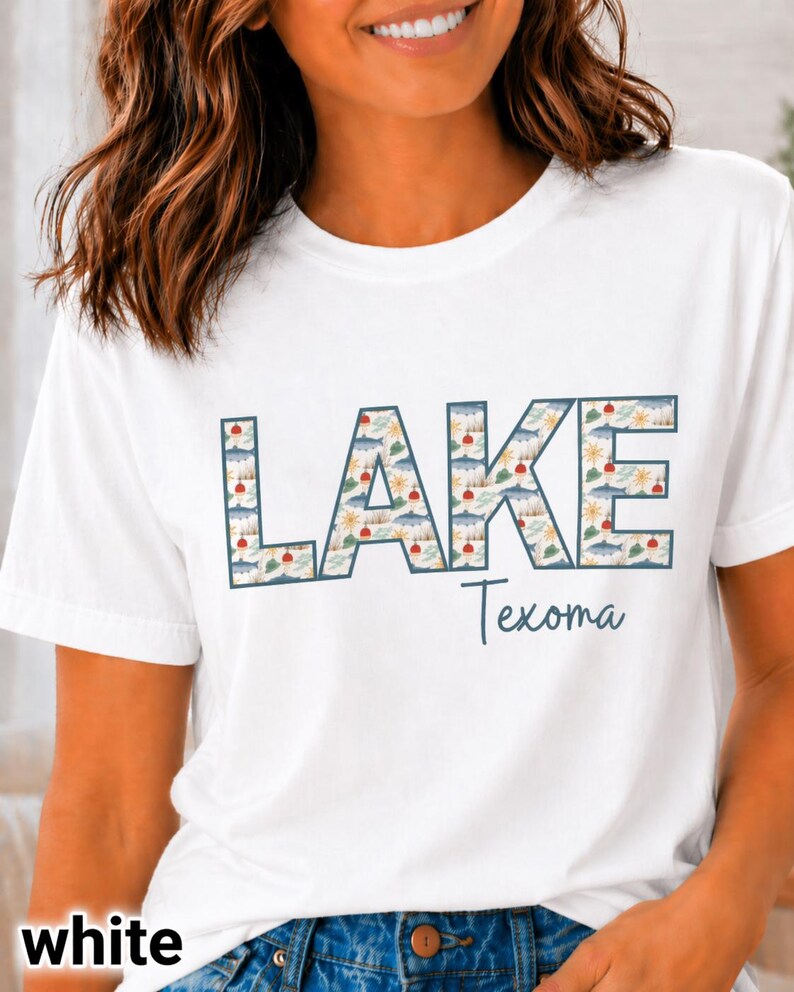 May include: White t-shirt with the word "LAKE" in large, patterned letters, and the word "Texoma" in script below. The "LAKE" letters feature a lake-themed design with clouds, suns, and fish.