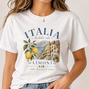 May include: White t-shirt featuring a watercolor design with the word "ITALIA" in blue, along with "la dolce vita" and "LIMONI". The design includes lemons, a coastal town, and the phrase "sole, limoni & mare".