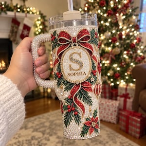 May include: A stainless steel tumbler with a handle, fully covered in small white crystals. It features a festive design with a large red bow, green pine branches, red berries, and a central circular emblem containing the letter S and the name SOPHIA.