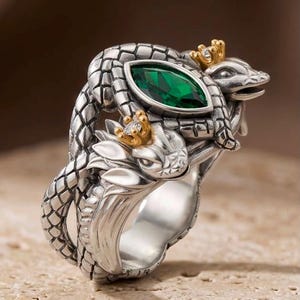 Puede incluir: Un anillo de plata con dos serpientes entrelazadas de escamas texturizadas. Cada cabeza de serpiente lleva una pequeña corona dorada con una diminuta piedra clara. Una gema verde esmeralda en talla marquesa está engastada en el centro.