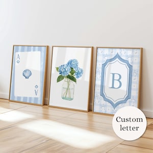 May include: Three framed wall art prints in light wood frames. The left print shows an ace of spades playing card with a seashell. The center print features blue hydrangea flowers in a glass jar. The right print displays a letter B in a frame.