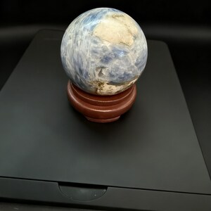 May include: A blue and white sphere, possibly a decorative stone or crystal, sits on a small, round, reddish-brown wooden stand. The sphere has a marbled appearance with shades of blue and white. The background is black.