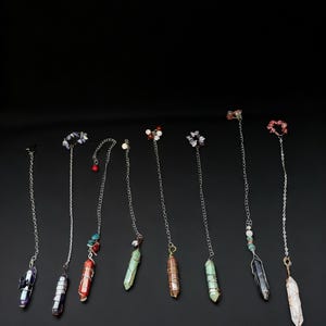 May include: A collection of crystal pendants with silver chains. Each pendant features a different colored crystal, including purple, red, green, and clear, with decorative beads at the top. The pendants are displayed against a black background.
