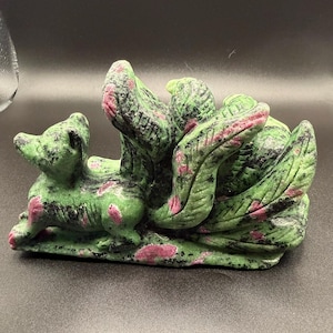 May include: A carved stone figurine of a multi-tailed fox made from green zoisite with embedded pink ruby inclusions and black mineral flecks. The fox is depicted in a reclining position with several curved, textured tails fanned out behind it.