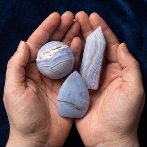 May include: Three blue lace agate stones in various shapes: a sphere, a teardrop, and a rectangular point. The stones are held in cupped hands against a dark blue background. The agate has white and blue bands.