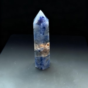 May include: A blue and white crystal obelisk with a pointed top. The crystal has a rectangular base and is standing upright on a dark surface. The crystal has a mix of blue and white colors with some brown and tan inclusions.