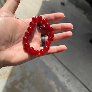 May include: A red beaded bracelet. The bracelet is made of round, polished red beads strung together on an elastic band. The beads are a deep, translucent red color. The bracelet is shown on a person's hand.