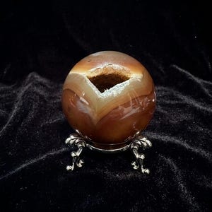 May include: A polished, spherical gemstone with brown and cream-colored bands, displayed on a decorative silver-colored stand. The sphere has a cavity in the center. The background is a dark, velvety fabric.