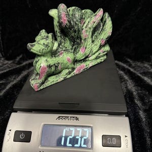 May include: A carved stone fox sculpture with nine tails, in shades of green, black, and pink, sits on a digital scale. The scale displays a weight of 1232 grams. The sculpture is set against a black background.