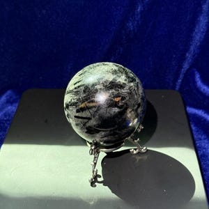 May include: A clear crystal ball with black inclusions, resting on a silver metal stand. The sphere is round and polished, reflecting light. The stand has a decorative, three-legged design. The background is a dark blue.