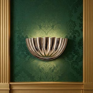 May include: A polished silver wall sconce with a scalloped design, mounted on a green damask wallpaper. The sconce is illuminated, casting a warm glow. The wall is framed by gold-colored trim and vertical accents.
