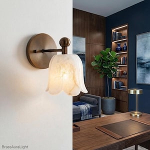 May include: A wall-mounted light fixture with a brass finish and a tulip-shaped, off-white shade. The fixture is mounted on a white wall, with a dark wood-paneled wall and a desk in the background. The text "BrassAuraLight" is visible.