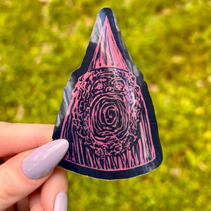 May include: A black sticker with a pink illustration of a creature with a cone-shaped hat. The creature has a wide open mouth and a spiral design in the center. The sticker is held against a blurred green background.