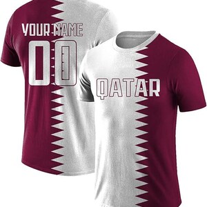 May include: Two white and maroon t-shirts. One shirt has "QATAR" printed on the front. The other shirt has "YOUR NAME" and the number "00" printed on the back. The shirts have a zig-zag design that splits the shirt in half.